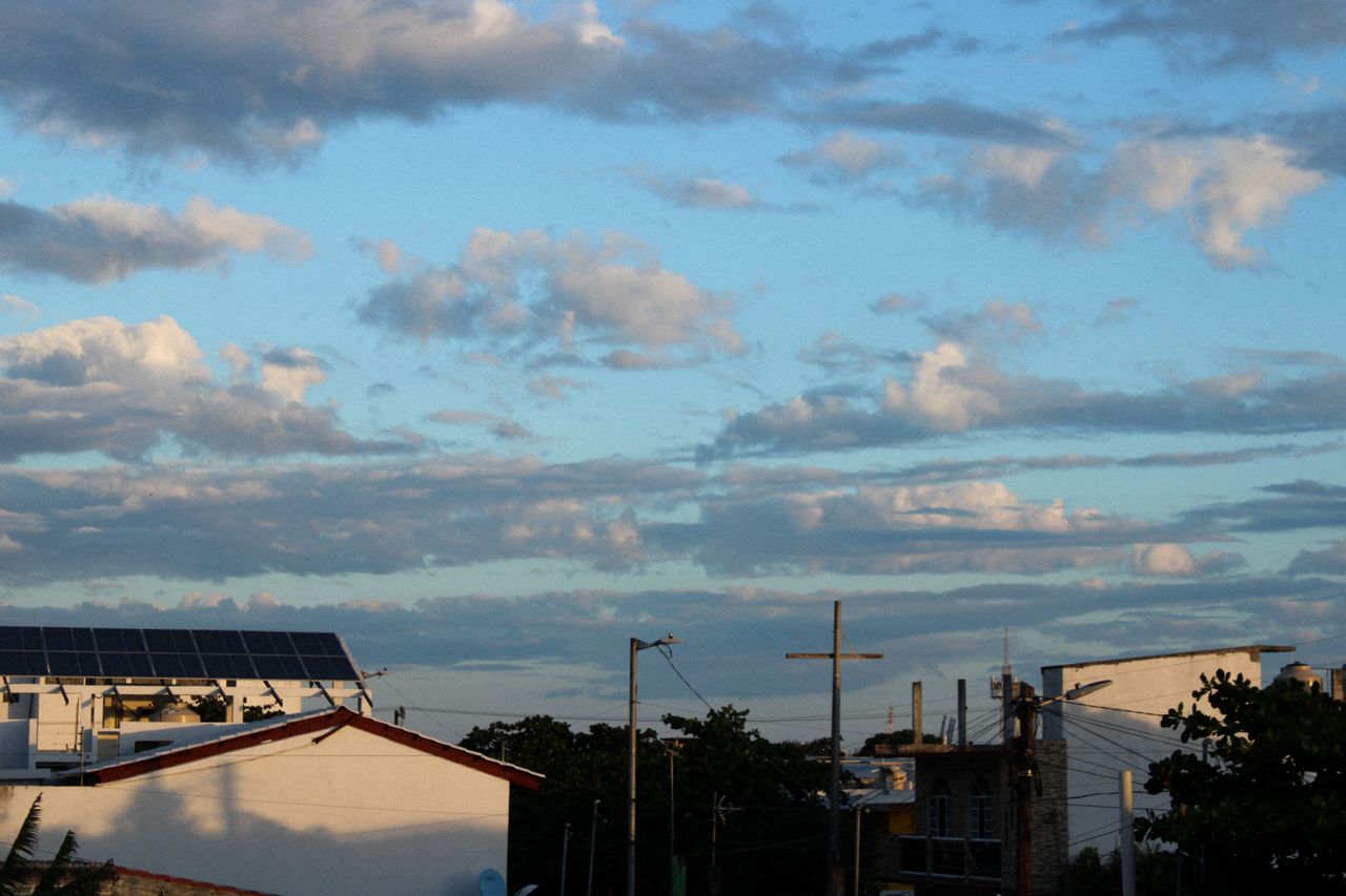 Services A scenic view of rooftops with solar panels under a cloudy blue sky during the day.
